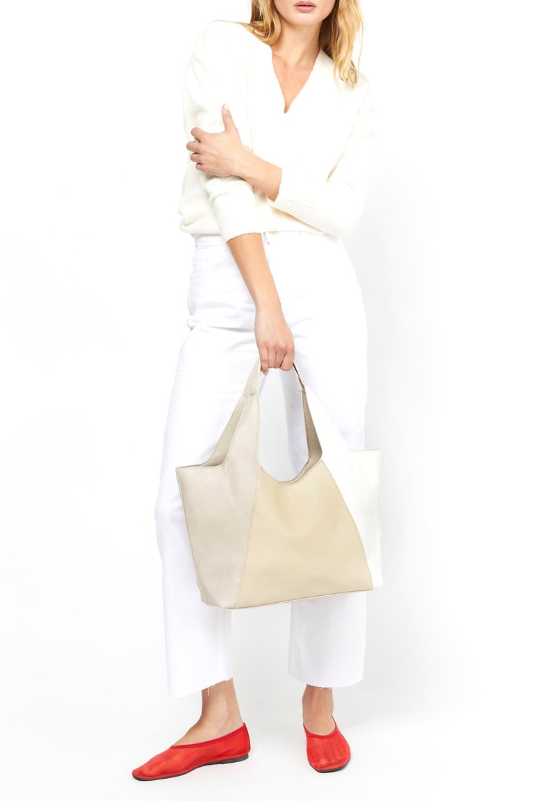 Beige läderhandväska med en slät yta, med kontrasterande paneler och robusta handtag, parat med vita byxor och röda ballerina skor.