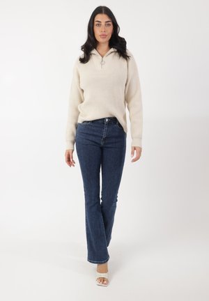 Pull beige en maille avec un col zippé moitié, associé à un jean flare bleu nuit. Le modèle porte des sandales à talons blancs ouvertes.