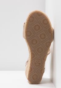 Světlý hnědý podrážka sandálu s texturovaným květinovým vzorem, vyrobená z přírodní gumy a s plochým tvarem základny. Viditelné logo UGG.