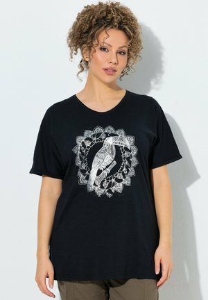 Donna con capelli ricci che indossa una maglietta nera con disegno bianco di tucano e mandala, in piedi davanti a uno sfondo chiaro semplice.