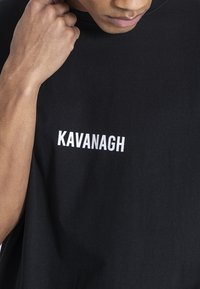 Černé bavlněné tričko s bílým potiskem loga "KAVANAGH". Materiál vypadá hladce, má krátké rukávy a kulatý výstřih.