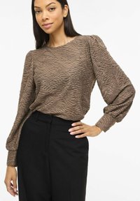 Blouse marron texturée avec motif ondulé, manches longues bouffantes, associée à un pantalon noir taille haute. Tissu lisse, coupe décontractée.
