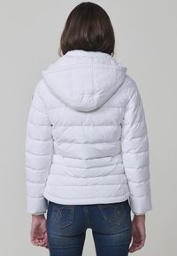 Chaqueta de plumas blanca con diseño acolchado horizontal, capucha y forro suave. Combinada con jeans azul oscuro que exhiben un sutil patrón bordado.
