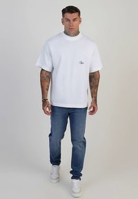 Camiseta blanca de algodón de gran tamaño con cuello redondo y un pequeño parche con logo. Combinada con jeans de mezclilla azul y zapatillas blancas.