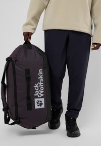Borsa a tracolla viola con accenti neri, caratterizzata da un grande logo e maniglie per il trasporto, appesa sulla spalla di una persona. Materiale nylon resistente.