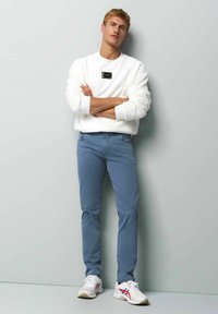 Sweatshirt blanc et fluffy avec un patch logo noir, associé à un pantalon slim-fit bleu clair et des baskets blanches avec des accents rouges et bleus.