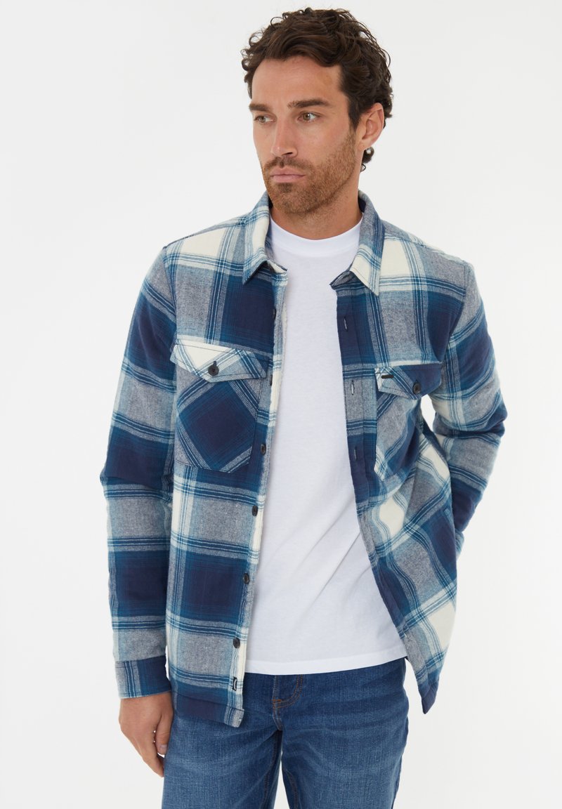 Blå og grå ternet flannel skjorte med to brystlommer, knappelukning foran og kontrasterende hvide detaljer; båret over en hvid T-shirt.