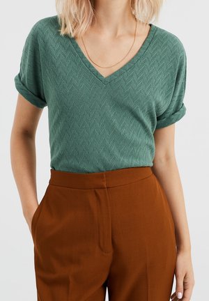 Kobieta nosząca zieloną teksturowaną bluzkę z krótkim rękawem i dekoltem w serek, włożoną w spodnie z wysokim stanem w kolorze rdzy, z cienkim złotym naszyjnikiem.