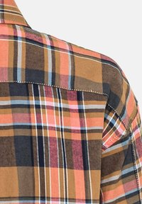 Gros plan d'un tissu de chemise à carreaux montrant un motif avec des lignes orange, marron, marine, rose et bleues et une couture visible sur la couture de l'épaule.