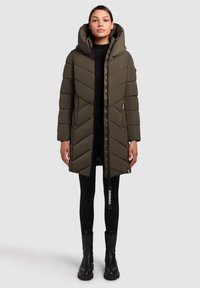 Oliivroheline puffjakker, millel on silmapaistev chevron-muster, varustatud kapuutsi, lukuga ja küljetaskutega. Kantud mustade retuuside ja saapade peal.