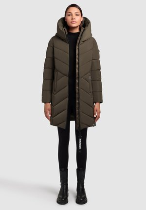 Olivgrüner Puffermantel mit ausgeprägtem Zickzackmuster, ausgestattet mit einer Kapuze, einem Reißverschluss und Seitentaschen. Getragen über schwarzen Leggings und Stiefeln.