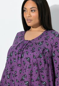 Fioletowa kwiecista bluzka z dekoltem w serek, zdobiona czarno-niebieskimi wzorami kwiatowymi, wykonana z miękkiego materiału, z marszczeniem na biuście dla dodatkowego efektu.
