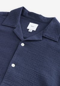 Tamnoplava košulja od teksturiranog tkanja, s ovratnikom i bijelim zakopčavanjem na gumbe. Unutrašnja etiketa na ovratniku nosi natpis "NEXT".