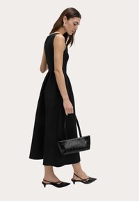 Schwarzes, ärmelloses Kleid mit tailliertem Oberteil und weit ausgestellt Rock. Kombiniert mit schwarzen spitzen Pumps und einer strukturierten schwarzen Handtasche.