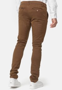 Brune slim-fit bukser med en struktureret finish, to baglommer med knapdetaljer, parret med hvide sneakers.