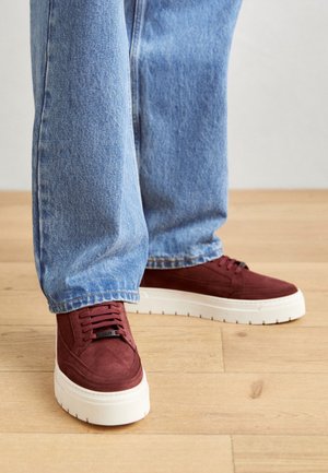 Persoon draagt lichtblauwe spijkerbroek en bordeauxrode suède sneakers met dikke witte zolen, staand op een houten vloer.