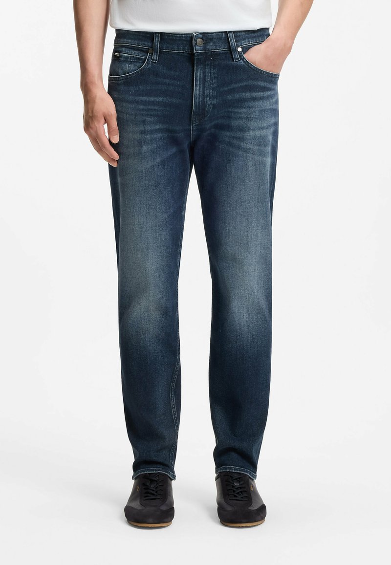 Homme portant un jean bleu délavé droit et des baskets noires, debout, une main dans la poche et l'autre détendue le long du corps.