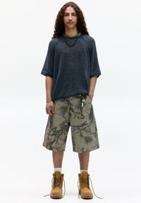 Cămașă scurtă cu mânecă, de culoare albastru închis și cu dungi, pantaloni scurți cargo bej cu un model camuflat și ghete de lucru bej. Accesoriile includ un colier și un breloc.