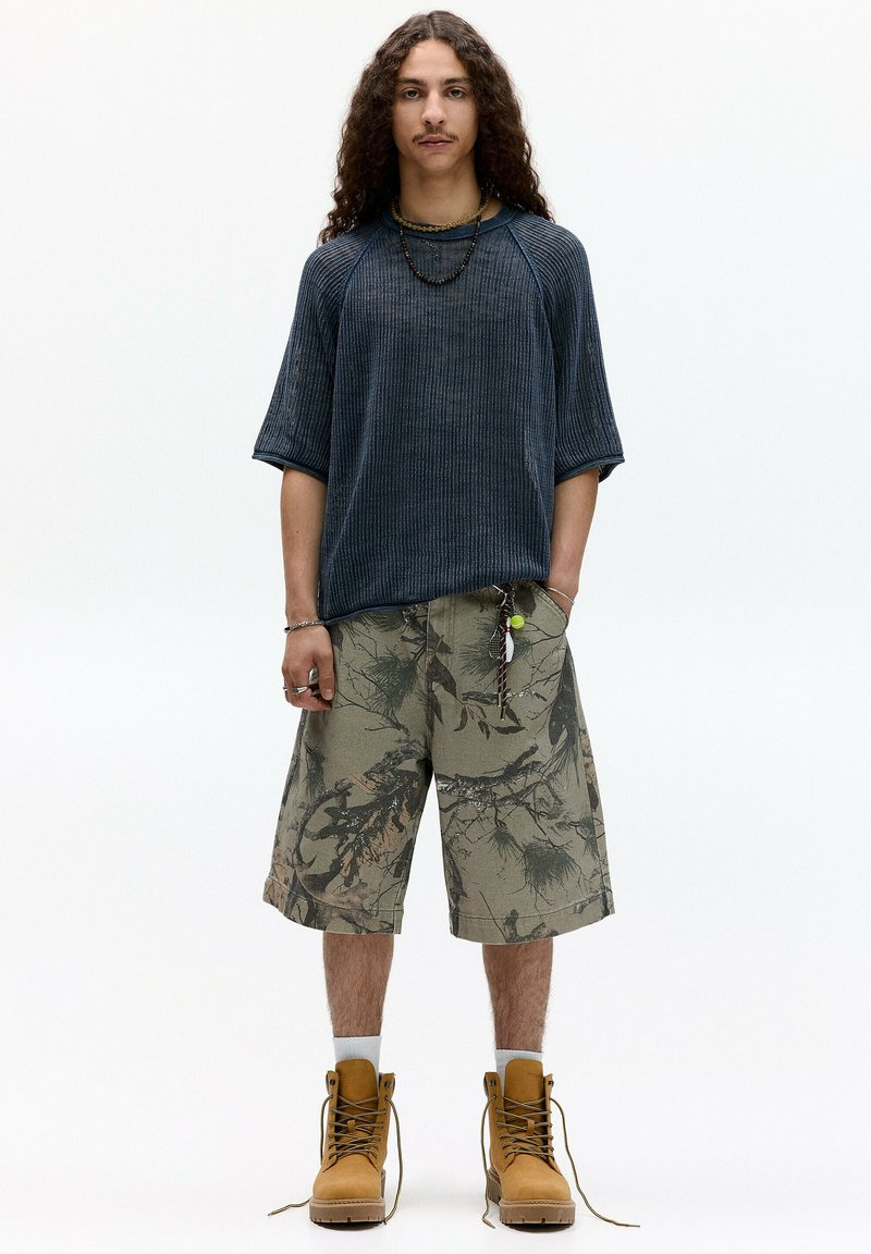 Dunkelblauses, gestreiftes Kurzarmhemd, tannfarbene cargo Shorts mit Camouflage-Muster und tannfarbene Arbeitsschuhe. Zubehör umfasst eine Halskette und einen Schlüsselanhänger.