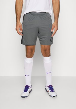 Mies, jolla on harmaat urheilushortsit, valkoiset polven korkeudelle ulottuvat sukat, joissa on violetteja logoja, ja valkoiset jalkapallokengät, joissa on violetit nauhat, seisoo valkoisella lattialla.