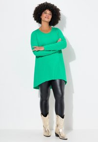 Grünfarbige Langarm-Tunika mit asymmetrischem Saum, kombiniert mit schwarzen Leggings und metallisch goldenen Cowboy-Stiefeln vor einem schlichten Hintergrund.