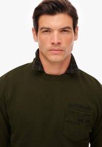 Sudadera de color verde oscuro con cuello redondo acanalado, que presenta un collar a cuadros y un bolsillo en el pecho con detalles de marca bordados.