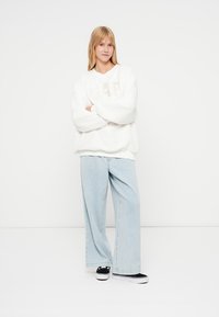 Felpa bianca in pile con tessuto testurizzato e logo beige, abbinata a jeans a gamba larga azzurri e sneakers nere.