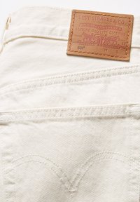 Jeans in denim bianco con un'etichetta in pelle marrone con testo impressa. Dettagli di cucitura visibili lungo le tasche e le cuciture.