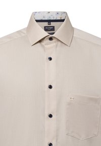 Beige textuur shirt met een klassieke kraag. Het heeft donkere knopen, een borstzak en een bloemenpatroon aan de binnenkant van de kraag.