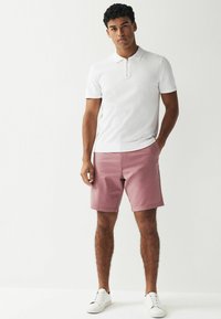 Hvid kortærmet polo med lynlås, parret med lyserød shorts. Modellen har hvide sneakers på. Glat stof, klassisk pasform, minimalistisk design.