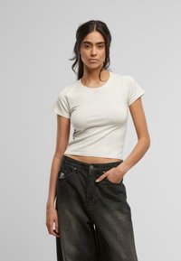 Kremfarget t-skjorte med korte ermer, tettsittende med rund hals, med et lite brodert logo på brystet, kombinert med svarte høye jeans.