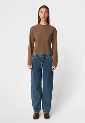 Kvinde stående iført en brun bluse med lange ærmer, løse blå jeans og mørkebrune sko mod en ensfarvet hvid baggrund.