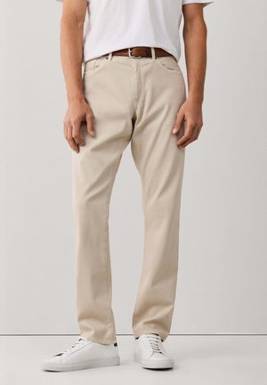Hombre que lleva pantalones beige de pierna recta, zapatillas blancas, un cinturón marrón y una camiseta blanca, de pie frente a un fondo liso.