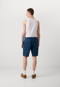 Shorts de cargo bleus avec une taille élastique, deux poches latérales et un détail d'étiquette. Portés avec un débardeur blanc et des chaussures brunes.