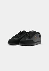 Black sneakers with smooth synthetic material, textured accents, lace-up design, and subtle gold branding on the side. Flat rubber sole.