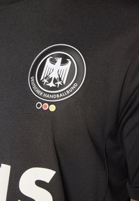 Melna džersija ar reljefu, uz kuras redzama apaļa emblēma ar divgalvainu ērglu un baltu tekstu “Deutscher Handballbund”.