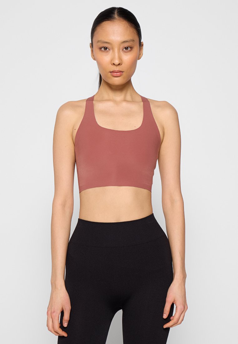 Eine Frau steht vor einem grauen Hintergrund und trägt ein ärmelloses, staubrosafarbenes Crop-Top und hochsitze schwarze Leggings. Sie schaut geradeaus mit einem neutralen Gesichtsausdruck.