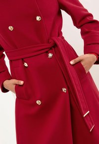 Un prim-plan al taliei unui model îmbrăcat cu o trench coat roșie, cu butoane decorative aurii și un cordon legat, având o mână în buzunar.
