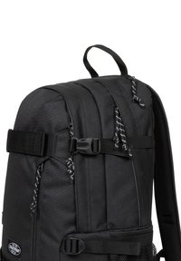 Mochila de tela negra con un diseño estructurado, que cuenta con correas ajustables, un bolsillo de malla y acentos en blanco a rayas en las cremalleras.