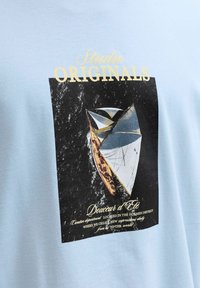 Voilier aux voiles blanches naviguant sur une eau sombre, surmonté du texte « Studio Originals » et de phrases décoratives en français sur un tissu bleu clair.