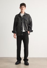 Chaqueta de mezclilla negra con cierre de botones, dos bolsillos en el pecho y mangas largas. Combinada con pantalones negros y zapatillas negras sobre un fondo neutro.