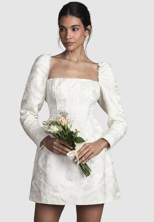 Donna in abito bianco a fantasia floreale con maniche lunghe a sbuffo che tiene un piccolo bouquet di fiori pastello legato con un nastro bianco.