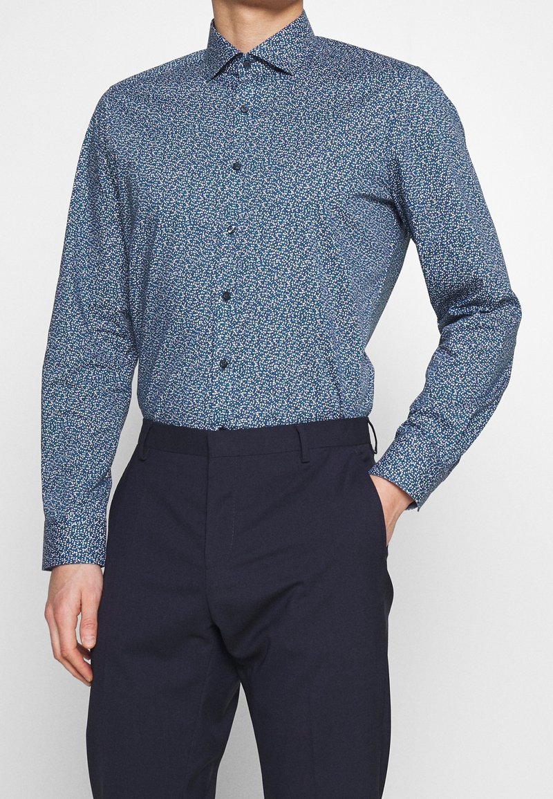 Homme portant une chemise cintrée à manches longues avec un petit motif floral blanc et bleu, associée à un pantalon bleu marine foncé.