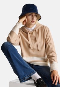 Haut à capuche beige et blanc à blocs de couleur avec un cordon de serrage, associé à un jean en denim évasé et un chapeau seau bleu marine avec un logo.
