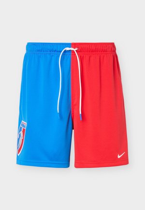 Mand iført blå og røde delte basketballshorts, hvide sokker, hvide sneakers og en marineblå hættetrøje med hænder afslappet langs siderne.
