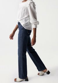 Weiße, gerippte Bluse mit rüschigem Ausschnitt und langen Ärmeln, kombiniert mit dunkelblauen, geraden Jeans und beigen, hohen Mules.