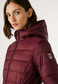 Vrouw met blauwe ogen in een bordeauxrood gewatteerd pufferjack met capuchon, zijkant logo patch en voorrits, tegen een effen achtergrond.