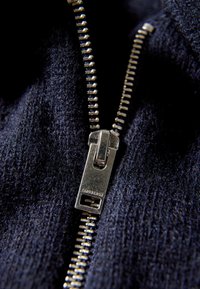 Granatowy materiał z zbliżeniem na metalowy zamek błyskawiczny z srebrną końcówką. Ząbki zamka są równomiernie rozmieszczone i widoczne na tle teksturowanego materiału.