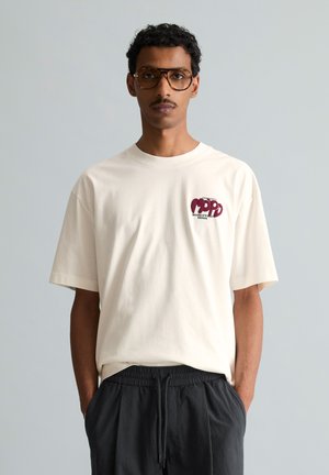 Uomo con capelli corti e ricci e occhiali, che indossa una maglietta bianca stampata e pantaloni neri con coulisse, in piedi davanti a uno sfondo grigio chiaro uniforme.