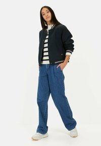 Marineblaue Bomberjacke mit gestreiftem Rippenkragen und -bündchen, über einem weiß-schwarz gestreiften Pullover getragen, kombiniert mit lockeren blauen Jeans und weißen Sneakers.